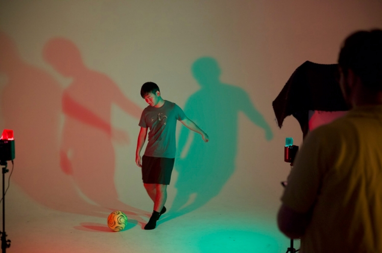 A Tisch Summer High School Photography student stands in front of a light portrait backdrop as he attempts to kick a soccer ball. The portrait studio is lit in soft neon light that casts large shadows of the student behind him.